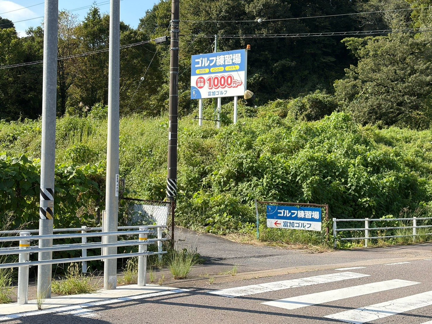 道路の横断歩道の先に見える「ゴルフ練習場 打ち放題1000円〜 富加ゴルフ」の大きな看板と、その手前の小さな案内看板